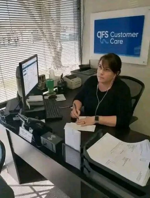 A woman wearing earphones sits at a desk with a computer monitor and documents, in front of a sign reading 'QFS Customer Care'.