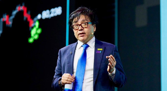 A man wearing glasses, a blue suit, and a bright blue tie speaks on stage, gesturing with his hand while holding a cup; behind him is a screen displaying a candlestick chart with green and red indicators and the number "80,238," alongside an "Endor" name badge. The image relates to the post by depicting Tom Lee, who is shown as the speaker referencing BitMine's recent $81M ETH purchase, tying his commentary to the bullish chart visible in the background.