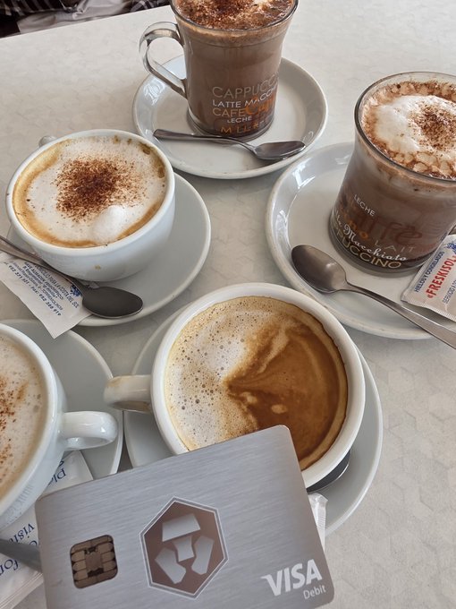 A table at a café displays several coffee drinks including cappuccinos and macchiatos, alongside a Visa Debit card and sugar packets.