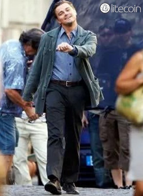 Leonardo DiCaprio walks and smiles during a film shoot, wearing a green jacket over a blue button-down shirt, with a "litecoin" logo visible in the top right corner of the image.