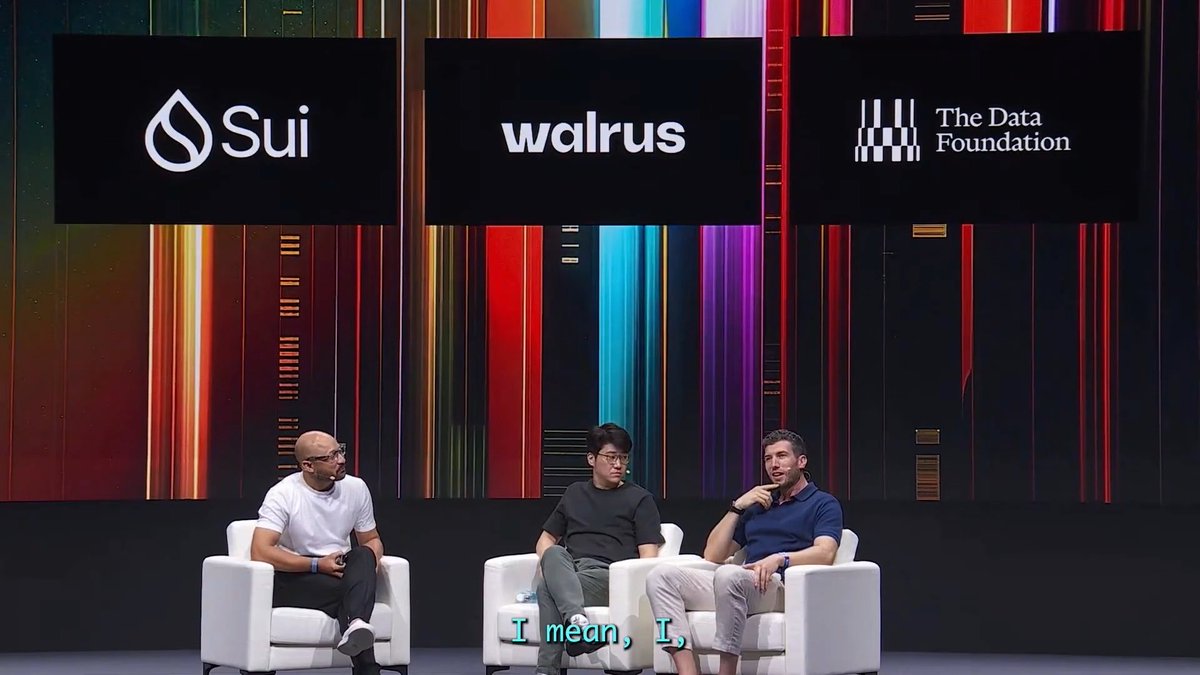 Three men sit in white chairs on a stage under large screens displaying logos for "Sui," "walrus," and "The Data Foundation," with the caption "I mean, I," visible at the bottom.