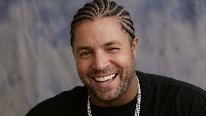 A portrait of a smiling man with braided hair and a beard, wearing a black shirt and pearl necklaces against a textured gray background.