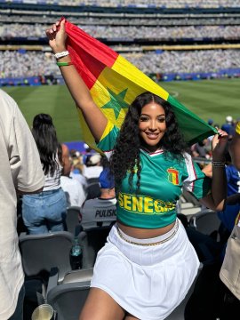 A Senegal fan wearing a green jersey with "SENEGAL" displayed across the front holds up her national flag at a stadium, celebrating her team's match against Belgium.