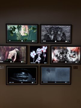 A gallery wall displaying seven framed screens or artworks arranged in a grid, featuring various images including flowers, a shirtless figure, white blossoms, and abstract black-and-white compositions.