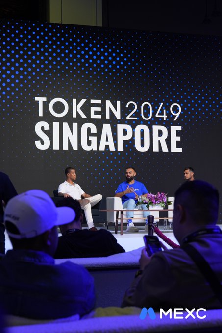 A panel discussion at the TOKEN2049 Singapore conference featuring three men seated on stage with the event branding and MEXC sponsor mark displayed prominently.