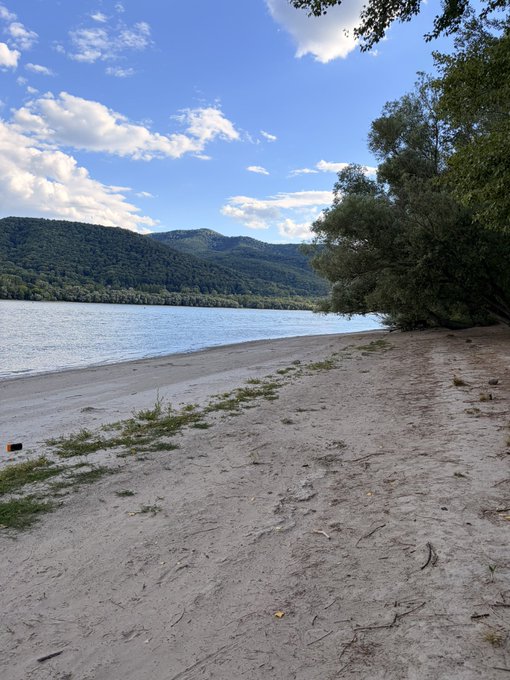 A scenic photograph of a sandy beach beside a calm blue body of water, with forested hills in the background and a partly cloudy sky.