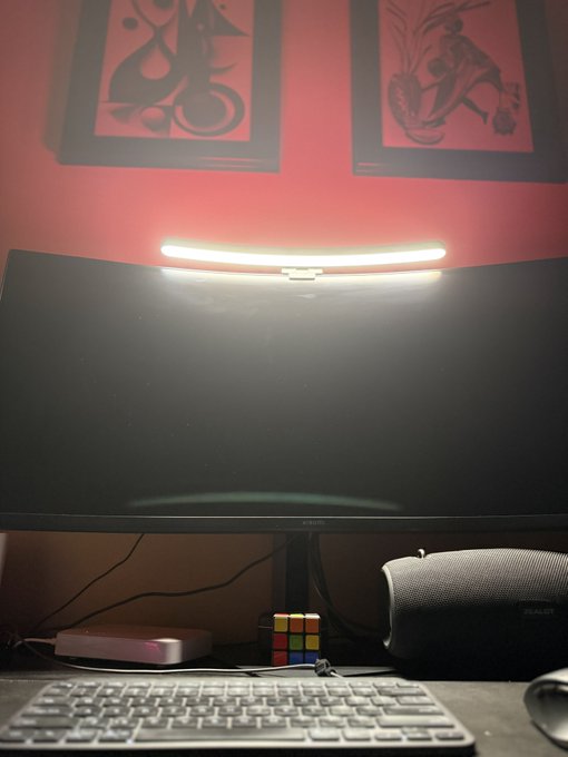 A desk setup featuring a curved monitor illuminated by a light bar mounted on top, with a colorful Rubik's cube, a keyboard, and two framed art prints on a red wall in the background.