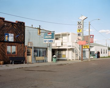 A small town street scene featuring old buildings including a 'Pepsi' and 'Pop Shoppe' storefront, with a tall vertical 'MOTEL' sign standing against a clear blue sky.
