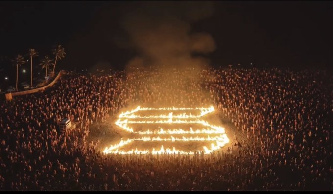 A large crowd gathers at night around a flaming logo in the shape of the Harley-Davidson bar and shield emblem, with palm trees visible in the background.