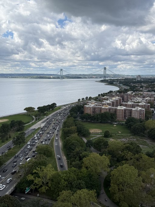 An elevated, wide-angle view shows a busy multi-lane highway curving along a coastline under a cloudy sky, with a large suspension bridge spanning the water in the distance.
