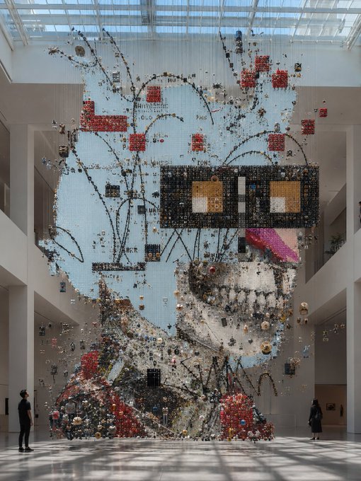 A massive suspended sculpture dominates a museum atrium, composed of thousands of small toy-like objects forming a face-like portrait with discernible text including "BALLISTIC" and "BLAIR." The scale of the installation is emphasized by two small visitors visible at the bottom, fitting the grand "Good Morning Legends" greeting and the celebratory #R3ORDR theme.