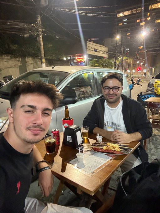 Two friends are enjoying a casual night out at an outdoor street-side table, sharing a plate of grilled skewers, drinks, and condiments while parked along a busy city street. The mention of @0xkaankacar suggests one of the men is the tagged friend, matching the relaxed social vibe of the photo.