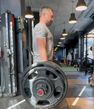 A man in a gray t-shirt standing in a gym, holding a large black weight plate, with exercise equipment visible in the background.