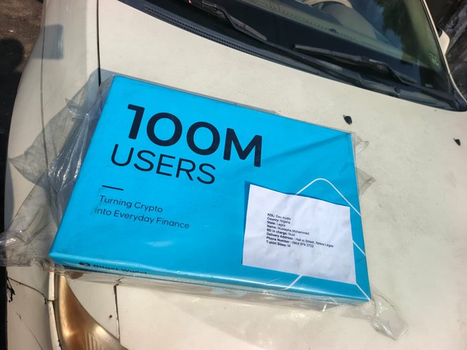 A package from Bitget Wallet, wrapped in plastic and featuring "100M Users - Turning Crypto into Everyday Finance" in bold black text on a blue cover, rests on the hood of a car, with a shipping label addressed to a KOL Lead in Uganda, celebrating the milestone of the wallet reaching 100 million users.
