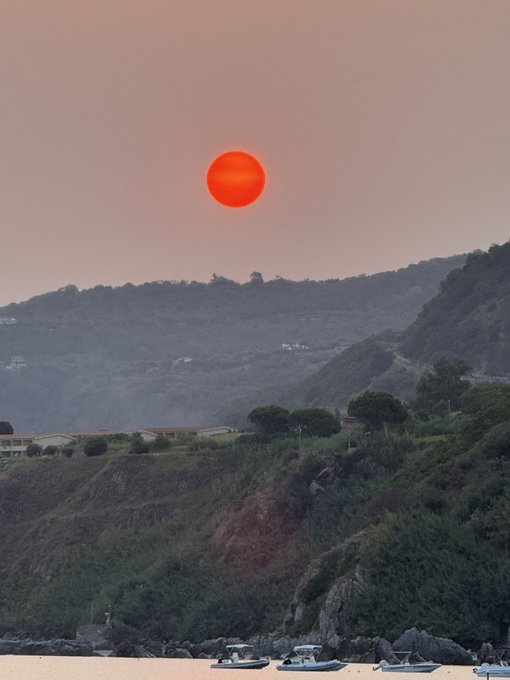 A scenic photograph of a red sun setting over hazy mountains and a coastal area with boats anchored in the water near rocky cliffs.