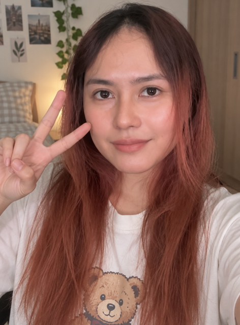 A young woman with long reddish-brown hair makes a peace sign while wearing a white t-shirt featuring a teddy bear graphic.