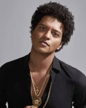A portrait of Bruno Mars wearing a black shirt and gold chain necklace with a medallion, looking at the camera against a plain background.