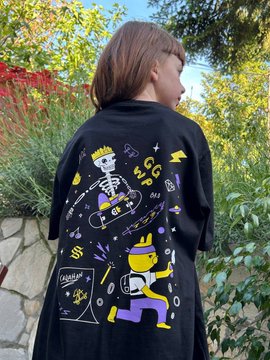 A young person with long hair wears an oversized black t-shirt featuring colorful cartoon-style graphics, including a skeleton, a skateboarder in a yellow helmet, and visible text like "GG MFG" and "SS," standing outdoors against lush greenery. The post celebrates young creative talent, with the shirt's hand-drawn, doodle-like design suggesting a DIY or streetwear aesthetic that fits the theme of showcasing up-and-coming artists.