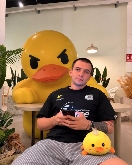 A man sits in front of a large yellow duck statue, holding a phone and with a small yellow chick plush toy on his lap, surrounded by plants in an indoor setting.