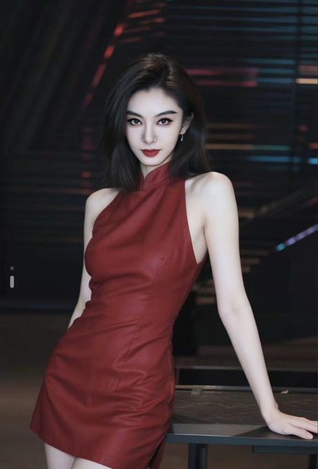 A woman with dark hair and red lipstick poses in a red leather halter dress at a dimly lit venue.