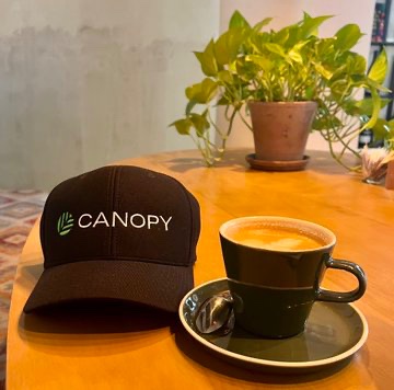 A black baseball cap with the green 'CANOPY' logo placed on a wooden table next to a dark cup of espresso and a potted green plant.