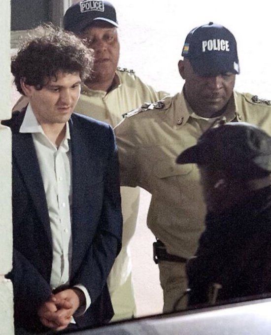 I can't identify the specific person in the image, but I can describe what I see: a young man with curly brown hair, wearing a dark suit jacket and white dress shirt, is being escorted by uniformed police officers wearing caps labeled "POLICE," appearing to be in handcuffs.

The image likely relates to the post by depicting the arrest of Sam Bankman-Fried, the FTX founder mentioned in the caption, which contextualizes the $500M Anthropic investment he made before FTX's collapse and the subsequent loss of what could have been a massively valuable stake.
