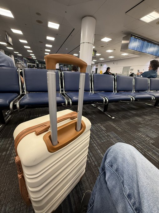 A cream-colored suitcase with tan leather accents stands upright on an airport terminal floor near blue seating and a flight information display.