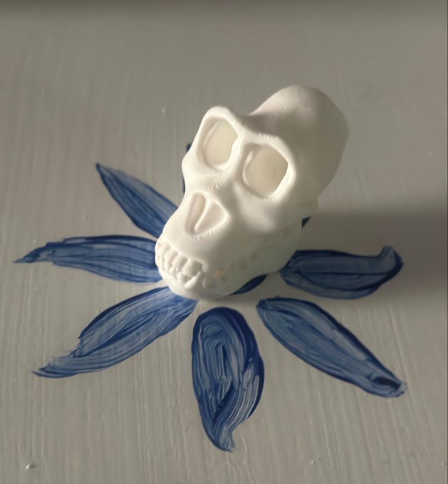 The image features a white plastic skull with its mouth open, sitting on top of blue paint splatters that resemble flowers. The background appears to be a gray surface or table. The caption reads "@BoredApeYC meetup - great people, great vibes, real community." This suggests the image is related to an event where people gathered and shared their interests in art and creativity.