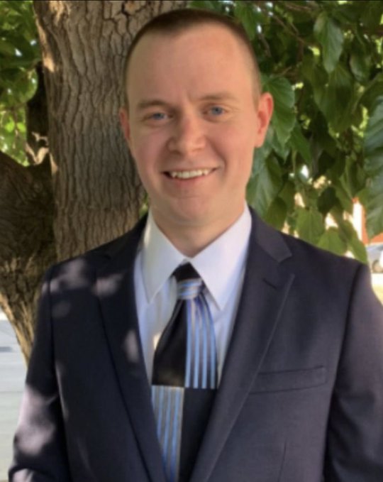 A portrait shows a smiling man with short hair wearing a navy blue suit jacket, white shirt, and blue striped tie, standing outdoors near a tree.
