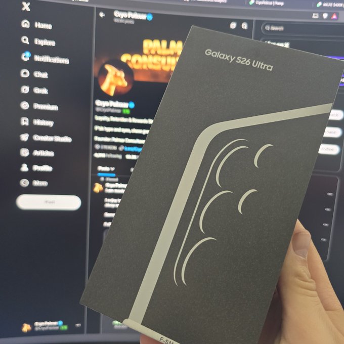 A hand holds up a black 'Galaxy S26 Ultra' smartphone box in front of a computer monitor displaying the X (Twitter) interface on the 'GypPalmer' profile.