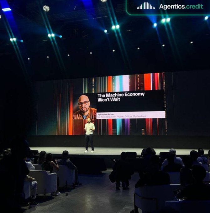 A speaker on stage presenting "The Machine Economy Won't Wait" at an Agentics.credit event, with his image on a large screen.