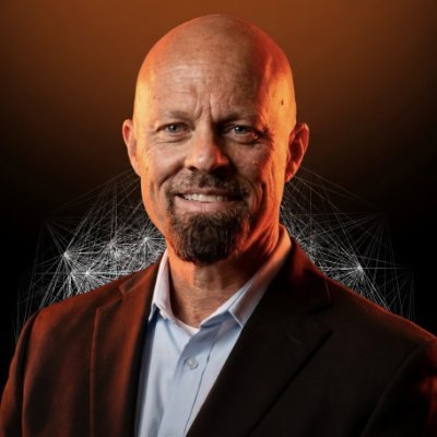 A portrait of a smiling bald man with a goatee wearing a dark suit and light blue shirt, set against a warm gradient background with a subtle geometric network pattern.
