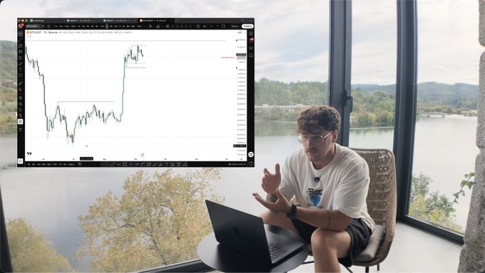 A young man sitting in a chair with a laptop by a large window overlooking a lake, with a YouTube video of a trading chart inset in the top left corner.