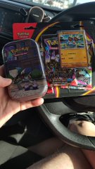 A hand holds up two Pokémon mini tins featuring artwork of Pokémon characters, including what appears to be Pikachu and other creatures, with Pokémon branding visible. In the background, additional Pokémon merchandise and a card are visible in a vehicle, corresponding to the post about opening a mini tin and getting two hits.