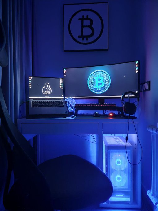 A dimly lit gaming/computer setup illuminated by blue LED lights, featuring a laptop and a curved monitor displaying Bitcoin logos, along with a PC tower with glowing blue fans and a framed Bitcoin sign on the wall.