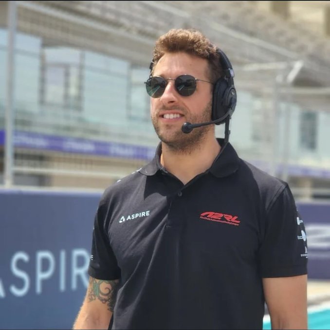 A man wearing sunglasses, headphones with a microphone, and a black polo shirt with 'ASPIRE' and 'TRL' logos stands outdoors, possibly at a racing venue.