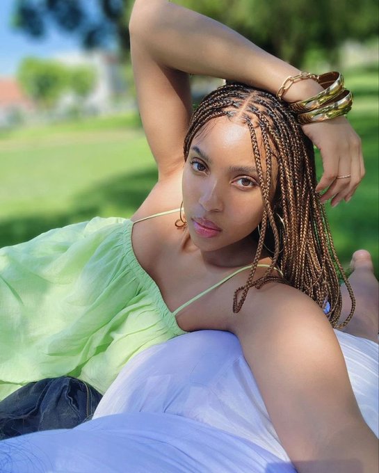 A portrait of a young woman with long braided hair, wearing a light green top and gold bangles, posing outdoors on a sunny day.