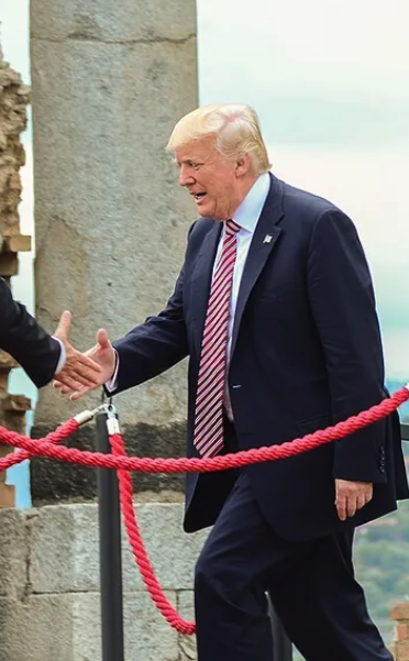 A photograph of Donald Trump wearing a dark suit and red striped tie, smiling and extending his hand for a handshake, standing near a red rope barrier.