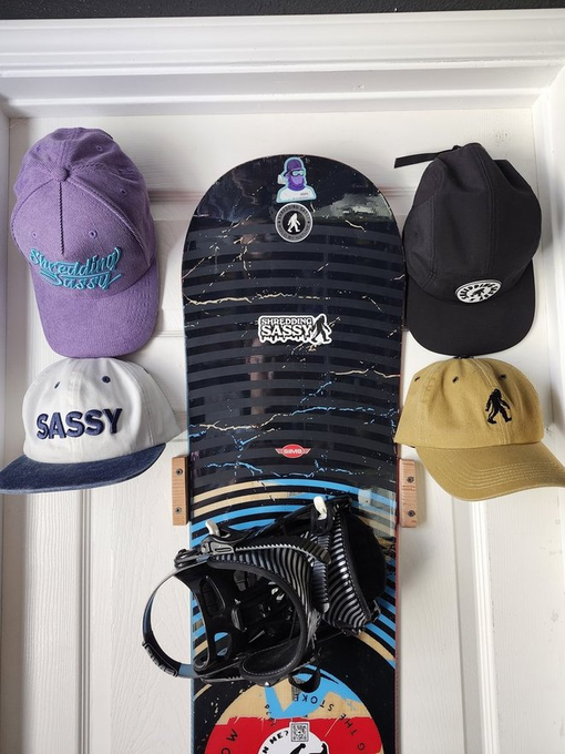 A snowboard mounted on a white door, surrounded by four caps labeled "Steadfast Sassy," "SASSY," and featuring astronaut and figure logos, with black bindings attached.