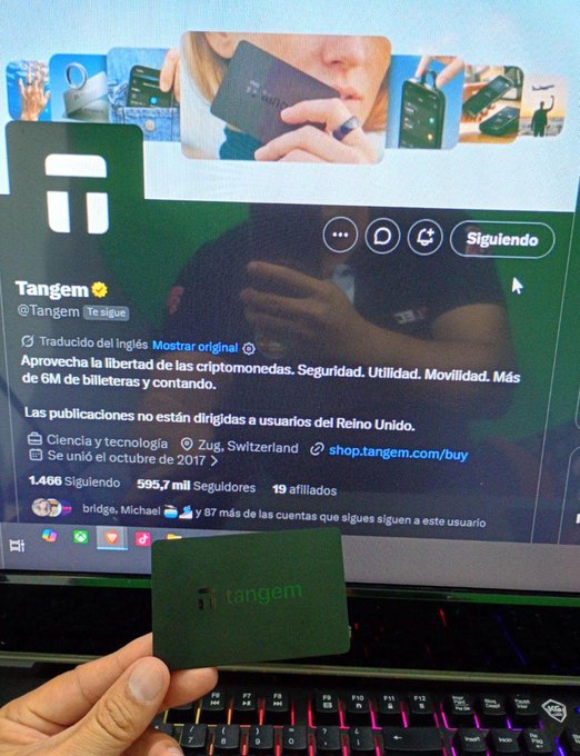 A hand holds a small green Tangem hardware wallet in front of a laptop screen displaying the Tangem Twitter/X profile, which describes it as a secure crypto wallet from Zug, Switzerland.