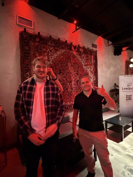 Two men pose in a dimly lit venue with red lighting; one wears a plaid shirt over a white t-shirt and the other wears a black polo and makes a hand gesture, standing in front of an ornate tapestry and a "Leadpool" banner.