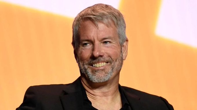 A portrait of Michael Saylor, the Bitcoin advocate and MicroStrategy founder, smiling in a black blazer against a yellow and white background.