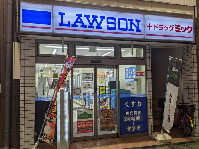 The image shows the storefront of a "LAWSON" convenience store with a "+ドラッグミック" (Drug Mic) pharmacy sign, featuring Japanese text advertising "くすり" (medicine) sales open 24 hours and Uber Eats delivery service. The image visually corresponds to the post about Lawson piloting JPYC stablecoin payments, as it depicts the Tokyo Lawson location where the cryptocurrency payment trial will be conducted in early August.
