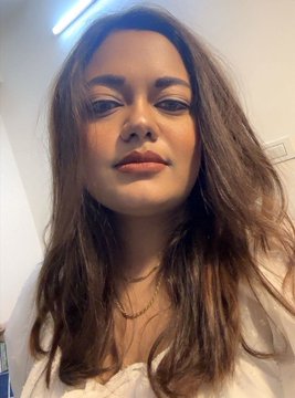 The image features a woman with long brown hair wearing a white shirt, looking directly at the camera while smiling. The background of the photo appears to be a bathroom or kitchen area, as there are two sinks visible in the scene.