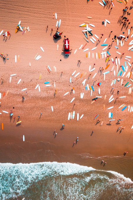 An aerial view of a beach crowded with surfboards scattered across the sand, a red rescue boat, and people walking near the shoreline where waves meet the shore.