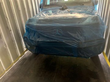 A photograph of a dark-colored car covered in a blue protective tarp, parked inside a metal shipping container.