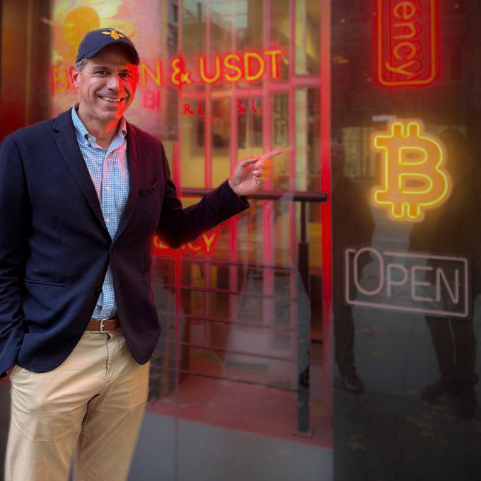 A smiling man wearing a navy blazer and a baseball cap with a bee logo points toward a window displaying neon signs reading "BITCOIN & USDT," an "OPEN" sign, and a glowing Bitcoin symbol. The image aligns with the post by showcasing HIVE's leadership in front of a Bitcoin-themed storefront, visually reinforcing the message of Bitcoin-powered, hydro-powered compute operations spanning four global cities.