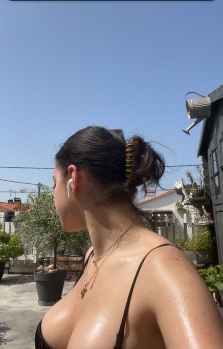 The image features a woman with her back turned towards the camera wearing a black bikini top, revealing her cleavage. She has a brown hair clip in her hair that matches the color of the sky behind her. The background includes a house and some potted plants, creating an outdoor setting for the photo. The caption to the image reads: "Last one on @dropee_app 🫩 Instead of chasing users, $DROPEE is building directly where they already exist. With Telegram’s integration into TON, that means access to over 1 billion users and transaction fees as low as $0.0005".