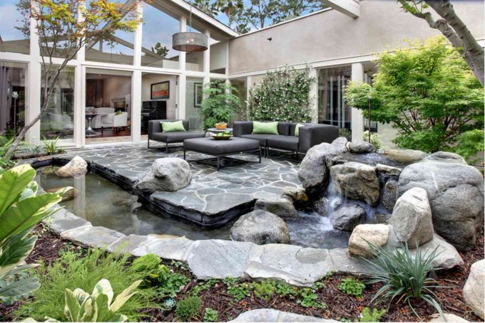 A serene outdoor patio features a stone-paved seating area with dark wicker furniture accented by green pillows, surrounded by a tranquil koi pond with a small waterfall, lush greenery, and modern home architecture.