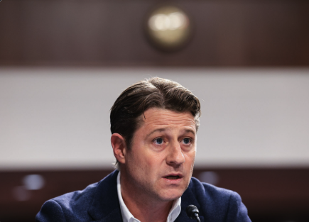 A man in a dark suit appears to be speaking at what looks like a formal hearing or testimony setting, with a serious expression on his face. The accompanying post humorously critiques Ben McKenzie for his skepticism toward Bitcoin's recent rise to $86,000, implying his previous bearish predictions have been proven wrong by the price surge.